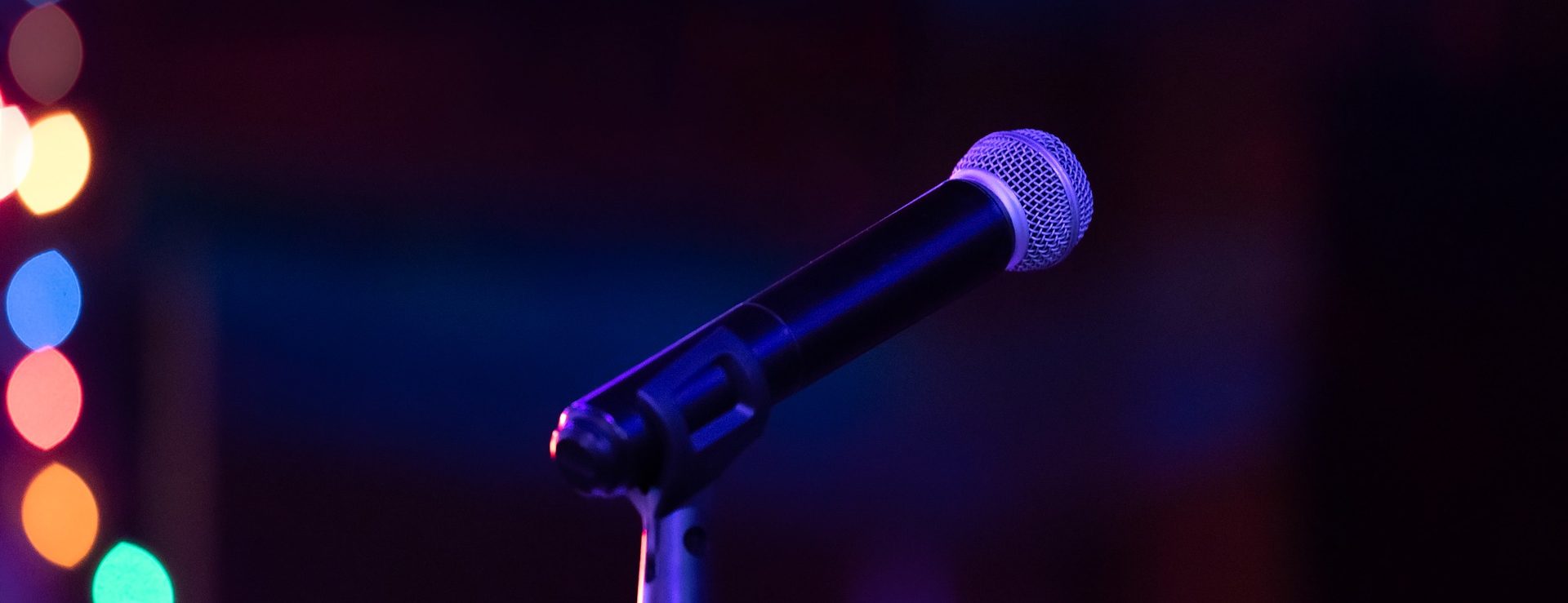 a dark background with multicolored lights blurred along the left side. A microphone on a stand is in the middle of the picture.