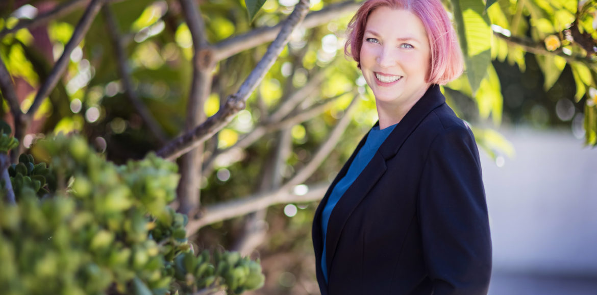 Picture shows an image of the coach and consultant, Kay Burnham, standing facing the left side of the screen with her head turned towards the reader. She is a white, female with pinkish red hair wearing a black blazer with a blue shell top. The background is outdoors in front of a tree where only a small grouping of branches and leaves is visible.